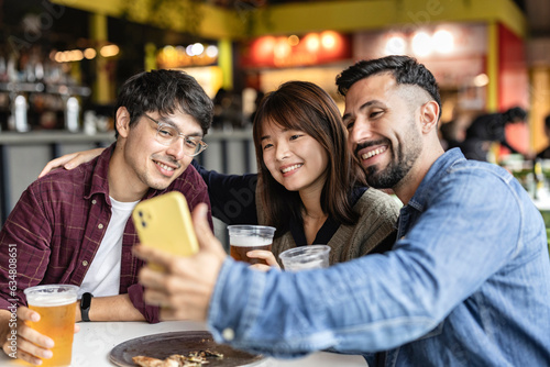 Multiracial friends toasting glasses of beer sitting in the brewery pub while taking a selfie with their smart phone