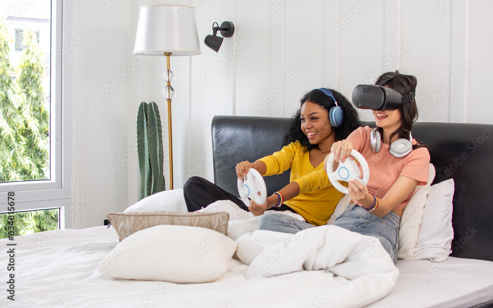 Portrait two diverse LGBT lesbian or female friends wearing casual clothes, sitting on bed in bedroom, holding game console and playing together with fun, smiling with happiness. Lifestyle Concept. Portrait two diverse LGBT lesbian or female friends wearing casual clothes, sitting on bed in bedroom, holding game console and playing together with fun, smiling with happiness. Lifestyle Concept.
