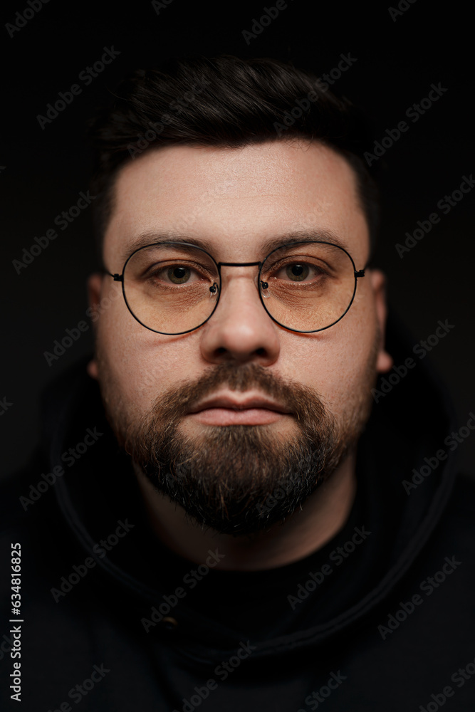 A close-up portrait of a bearded man wearing glasses and a black hoodie. He is looking at the camera with a serious expression. The background is black A close-up portrait of a bearded man wearing glasses and a black hoodie. He is looking at the camera with a serious expression. The background is black