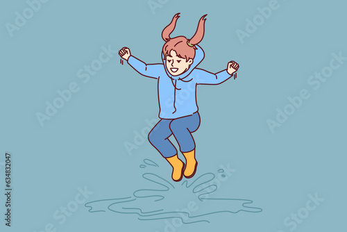 Little girl jumps in puddles in rubber boots and enjoys rainy autumn weather. Laughing child dressed in raincoat plays and bounces in puddles and has fun rejoicing in walk in fresh air