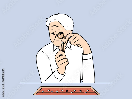 Elderly man jeweler examines diamond through magnifying glass checking gems for authenticity. Gray-haired worker in jewelry workshop chooses diamond to create necklace or engagement ring.