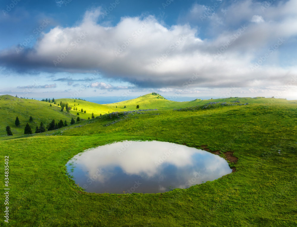 Obraz premium Aerial view of pond and green alpine meadows and hills at sunset in summer. Top drone view of mountain valley, green grass and sky with clouds reflected in water. Velika Planina, Slovenia. Landscape