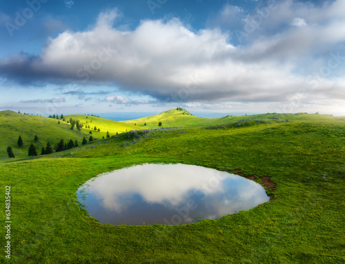 Aerial view of pond and green alpine meadows and hills at sunset in summer. Top drone view of mountain valley  green grass and sky with clouds reflected in water. Velika Planina  Slovenia. Landscape