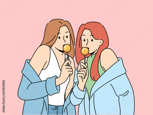 Two women with chupa chups in hands dressed in fashionable seductive clothes eat lollipops. Lovely girls in stylish jackets slung off shoulders to advertise new candies for teenagers