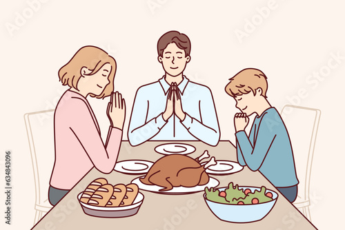 Family prayer before dinner, sitting at table with delicious food and thank god for well-being. Happy religious man and woman together with son say prayer, observing catholic ritual.