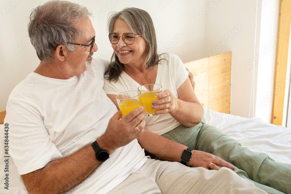 Cheerful old european husband and wife have breakfast, cheers, enjoy glasses of juice, sit on bed Cheerful old european husband and wife have breakfast, cheers, enjoy glasses of juice, sit on bed