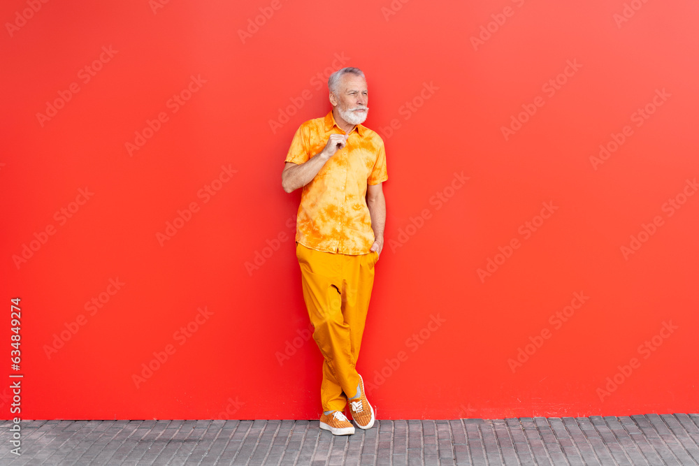 Handsome serious senior man, bearded hipster wearing stylish orange clothes waiting, standing on the street, isolated on red background. Fashion model posing for pictures outdoors, looking away