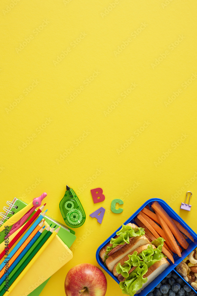 Delicious and nutritious school break. Top view vertical photo of educational stationery, sandwiches and organic treats on yellow background with blank space for promo or message