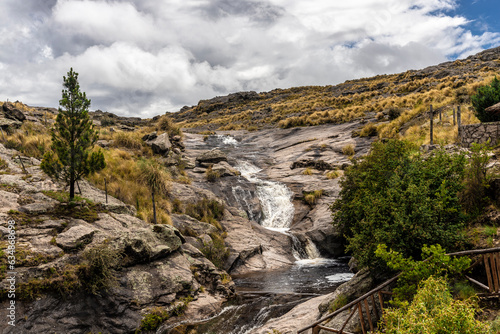 Salvaje cascada de agua natural en medio de una montaña de rocas, rodeada por pastos secos de monte y árboles. Camino a las Altas Cumbres, Córdoba, Argentina