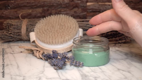 Women hand checking consistency of natural homemade face cream in transparent jar, marble white background with dark wood and dry lavender flowers.