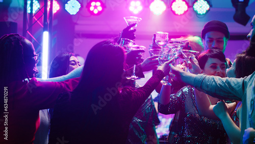 Happy diverse people clinking glasses at disco party, drinking alcohol and saying cheers on dance floor. Men and women partying and having fun at nightclub, raising drinks at event. photo