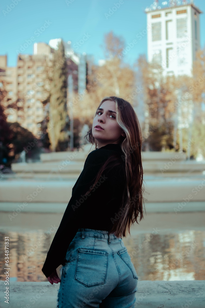 portrait of young latin woman from behind looking up dressed in jean and black t-shirt in a park with fountain behind in autumn portrait of young latin woman from behind looking up dressed in jean and black t-shirt in a park with fountain behind in autumn