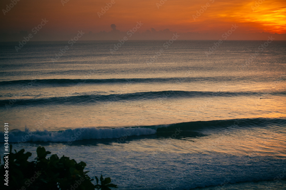 Viewed from above, a wave rolls gracefully, its crest illuminated by the sun's amber descent, casting a golden silhouette against the deepening hues of the horizon.