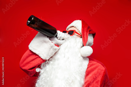 Foto drunk santa claus in hat and festive glasses drinks wine from bottle on red back
