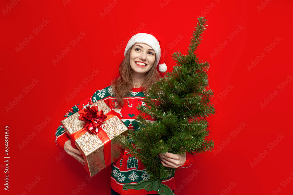 young cute girl in christmas sweater and santa hat holds gift and christmas tree on red background