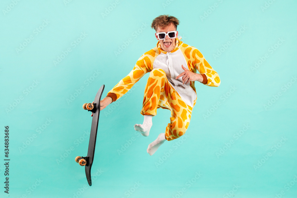 young funny guy in orange giraffe pajamas jumps and flies with skateboard on blue isolated background