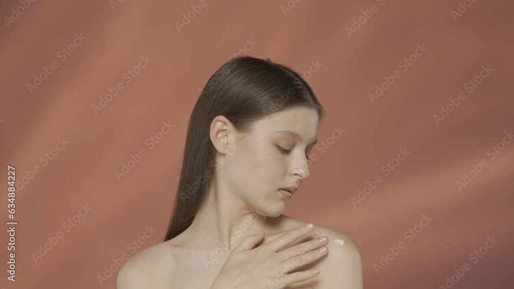 Attractive seminude woman applies, rubs body cream into the skin of her shoulders. Young woman in the studio on a red background. The concept of beauty, cosmetology, care. HDR BT2020 HLG Material.