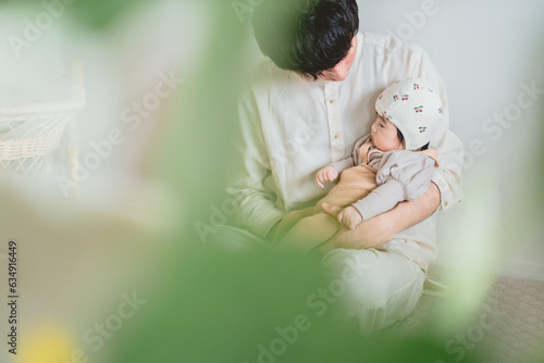 秋の日中,部屋の中で座って膝の上に子どもを抱っこするお父さんと、腕の中で眠そうにしている帽子を被った日本人の赤ちゃん photo