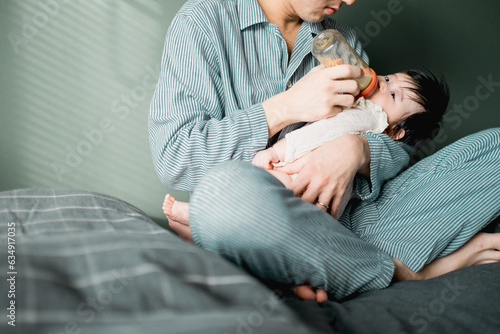 春の朝、布団の上で赤ちゃんに哺乳瓶でミルクを飲ませるのパジャマ姿のアジア人の男性