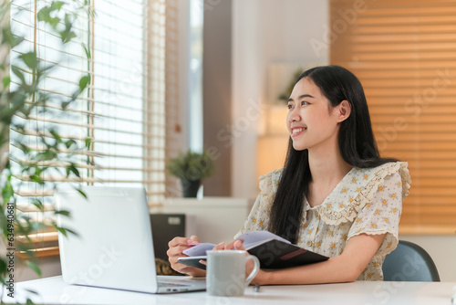 Smiling female entrepreneur taking notes, making business plans on notebook and using laptop at workplace.