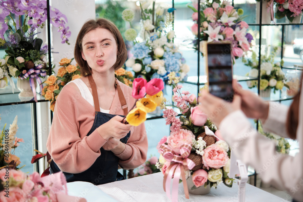 Fototapeta premium E-commerce business, one young White female florist demonstrates and shows floral arrangements via online live streaming with smartphone application in bright flower shop, beautiful blossoms store.