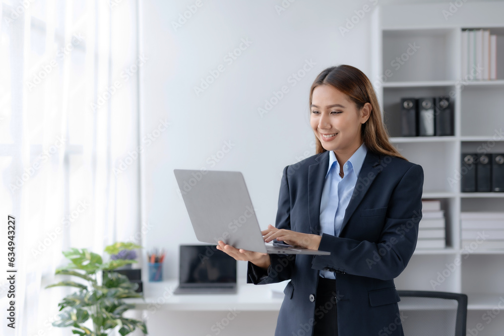Successful Asian businesswoman smiling stand using laptop computer at office. Confident Asia businesswoman standing happily in the office.