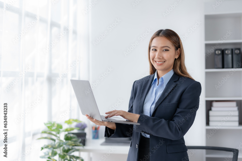 Successful Asian businesswoman smiling stand using laptop computer at office. Confident Asia businesswoman standing happily in the office. Successful Asian businesswoman smiling stand using laptop computer at office. Confident Asia businesswoman standing happily in the office.
