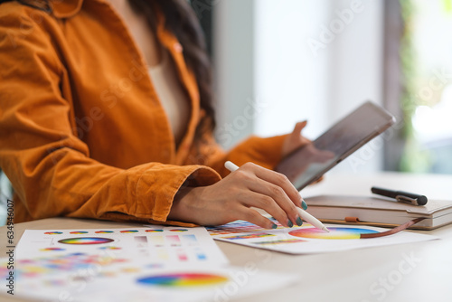 Cropped shot of female designer holding digital tablet, working with color samples at desk.