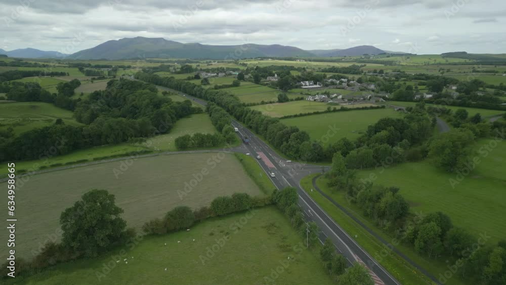 Busy rural road A66 with mountain Blencathra in the distance and green patchwork fields on overcast summer day. English Lake District, Cumbria, UK.