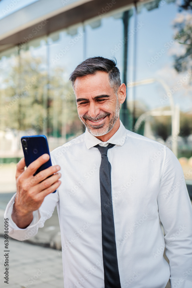 Vertical portrait of a mature businessman using a cellphone to to read job offers online. Successful corporate man watching good news with smartphone. Executive male winning with his stock shares Vertical portrait of a mature businessman using a cellphone to to read job offers online. Successful corporate man watching good news with smartphone. Executive male winning with his stock shares