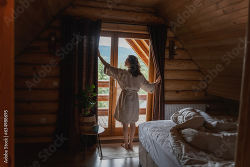 Young caucasian woman opening curtains in a wooden chalet cabin in the mountains.