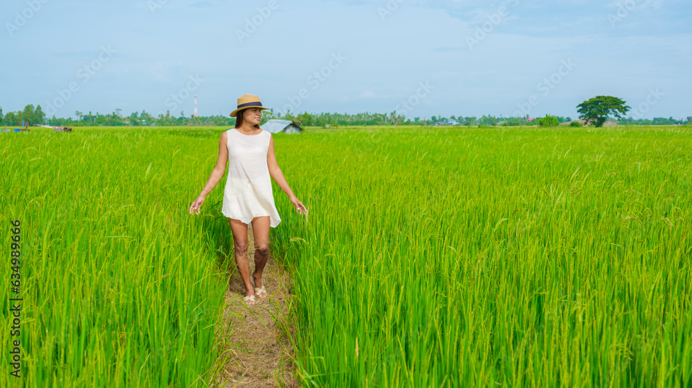 Aian woman on vacation in Thailand walking on a countryside road in the middle of green rice paddy fields in Central Thailand Suphanburi region, drone aerial view of green rice fields in Thailand
