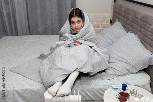 Foto Sick young girl wraps herself in a blanket and warms up at the heater
