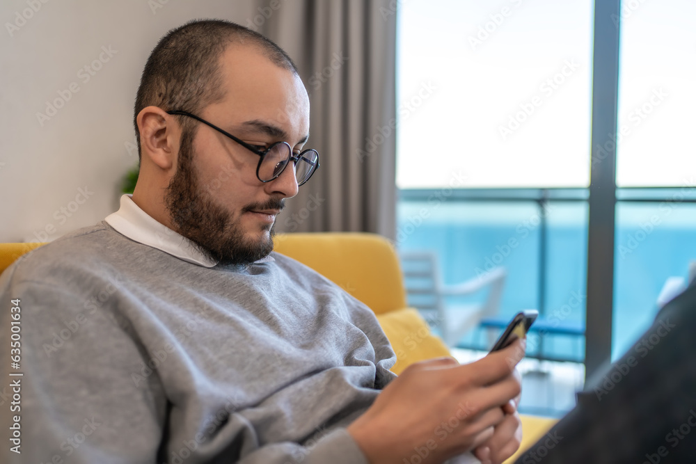Man sitting on couch looks at mobile phone. Guy checking email on smartphone or reading news in social media. Man sitting on couch looks at mobile phone. Guy checking email on smartphone or reading news in social media.