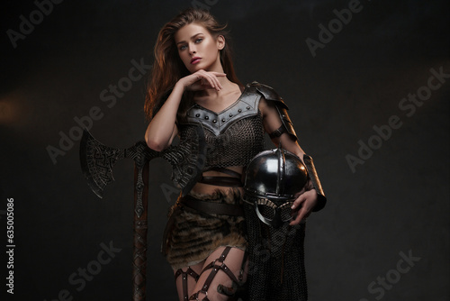 Beautiful Viking warrior model posing with a powerful axe and helmet showcasing strength and femininity in a medieval-inspired costume against a textured background © Fxquadro