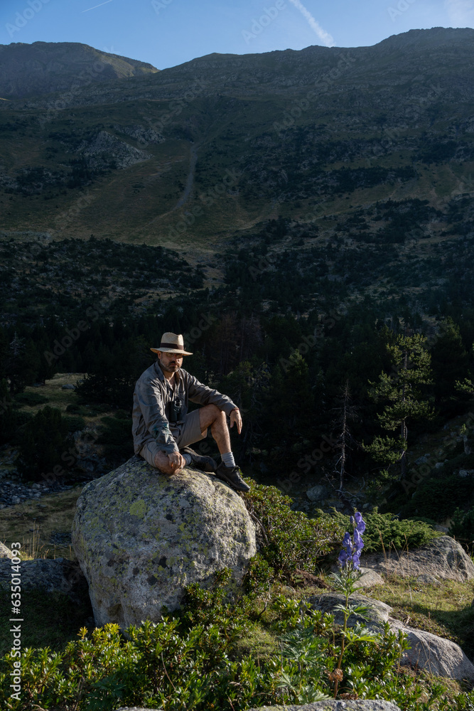 man sitting on large rock posing and looking at camera in mountain scenery, dressed in khaki with straw hat and scout-like appearance man sitting on large rock posing and looking at camera in mountain scenery, dressed in khaki with straw hat and scout-like appearance