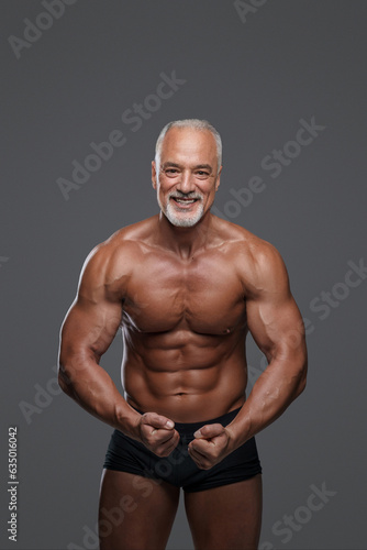 Charming mature muscular male model with stylish gray beard, wearing black underwear and posing with a big smile against a gray background