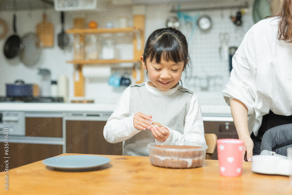 春の日中にキッチン前のテーブルで笑顔でケーキ作りをする日本人小学生の女の子と母親