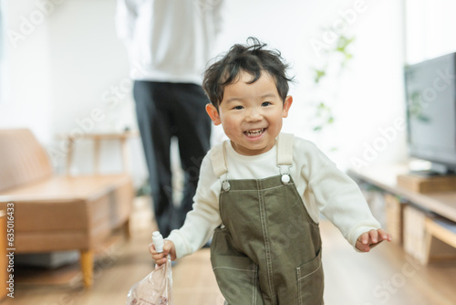 春の日中のリビングでおもちゃを持って笑顔で走るアジア人の息子と後ろから見守る父親 photo