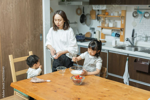 秋の日中の家の台所でお菓子作りをするアジア人の小学生とその手伝いをする母親と弟