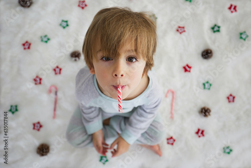 Little boy in pajama sitting on the floor on a white carpet with Christmas decorations.