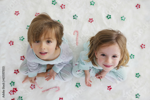 Blond twins in pajamas sitting on the floor on a white carpet with Christmas decorations.