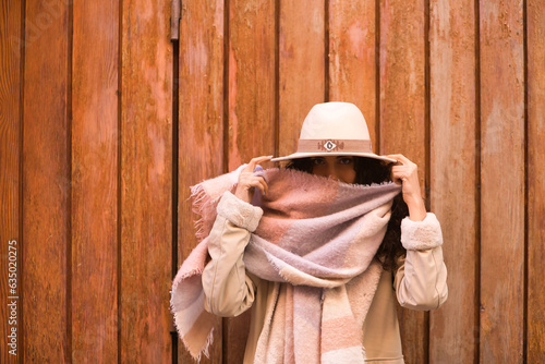 Young, pretty, brunette woman with curly hair and coat, scarf and hat, looking at camera while playing funny with her scarf, on wooden background. Concept beauty, fashion, autumn, winter, cold.