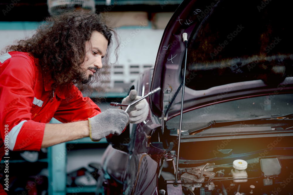 Long hair handsome mechanic man in red uniform works with engine vehicle, auto mechanic technician inspecting maintenance engine system of customer car automobile at garage car repair service shop.