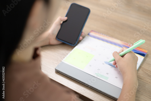 A woman checking planner on mobile phone with taking notes on calendar. Take notes for meeting appointments or Debt payment. Event planner timetable agenda plan. Calendar event plan, work planning
