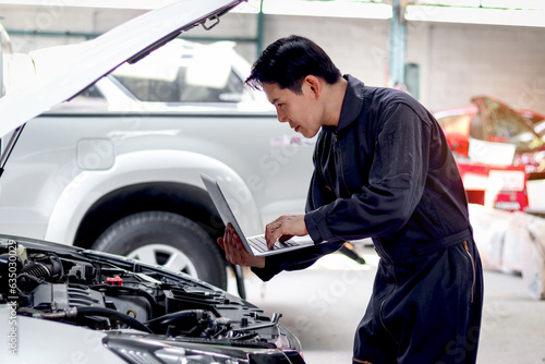 Asian mechanic man in uniform holding laptop computer during working with engine vehicle, auto mechanic technician inspecting maintenance customer car automobile at garage car repair service workshop.
