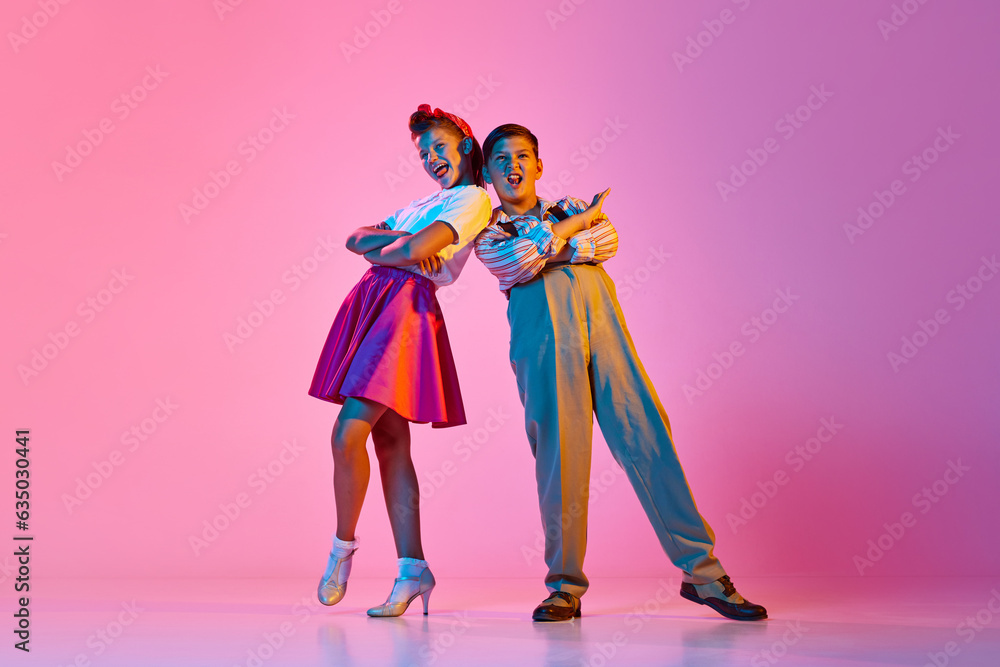 Stylish children, boy and girl on retro style clothes posing with emotive face, facing against pink studio background in neon light. Concept of childhood, hobby, active lifestyle, performance, art