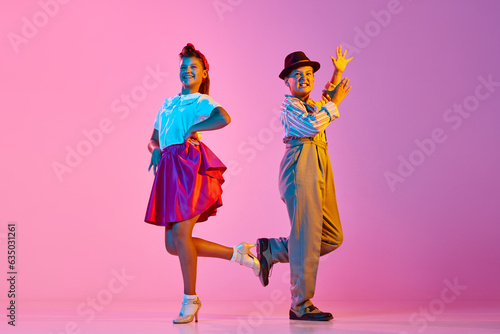 Emotional, beautiful children, boy and girl in retro costumes dancing lindy hop against pink studio background in neon light. Concept of childhood, hobby, active lifestyle, performance, art, fashion