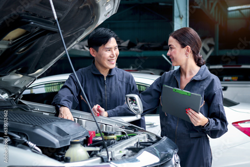 Happy mechanic man and woman mechanic in uniform discussing while working together with engine vehicle at garage, two auto mechanic technician repairing customer car at automobile repair service shop.