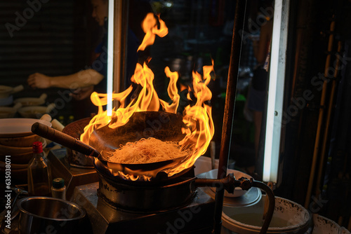 Photography Close-up of a wok frying noodles over an open flame at a Thai night market